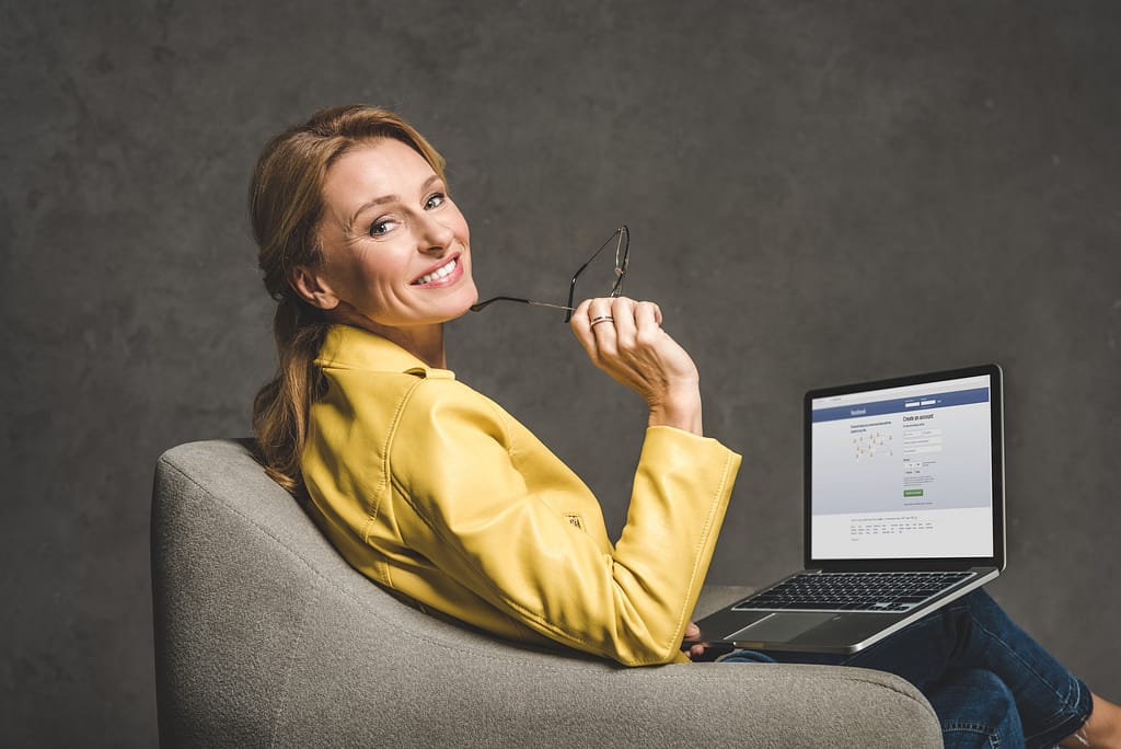 smiling mature woman using laptop with facebook website and sitting in armchair